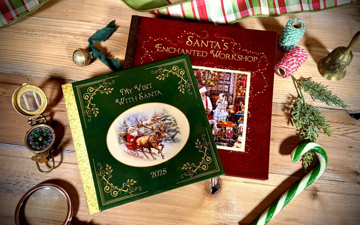 Two custom keepsake storybooks — a green “My Visit With Santa 2025” and a red “Santa’s Enchanted Workshop” — on the workshop table